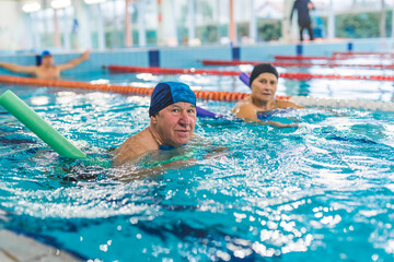 active elderly couple learning to swim with the help of swim noodles, indoor public pool. High quality photo