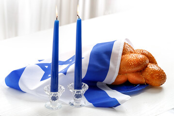 Challah bread covered with the Israeli flag, burning blue candles against a white background. Traditional Jewish Shabbat ritual. Shabbat Shalom.