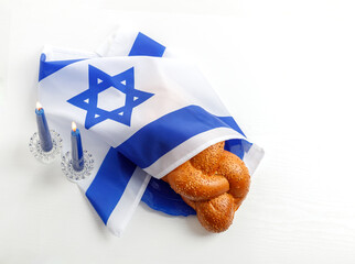 Challah bread covered with the Israeli flag, burning blue candles against a white background. Traditional Jewish Shabbat ritual. Shabbat Shalom.