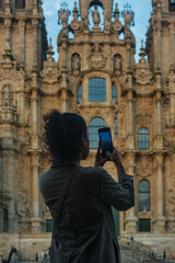 Young Hispanic woman with curly hair taking a photograph with her mobile phone of the facade of the Cathedral of Santiago de Compostela