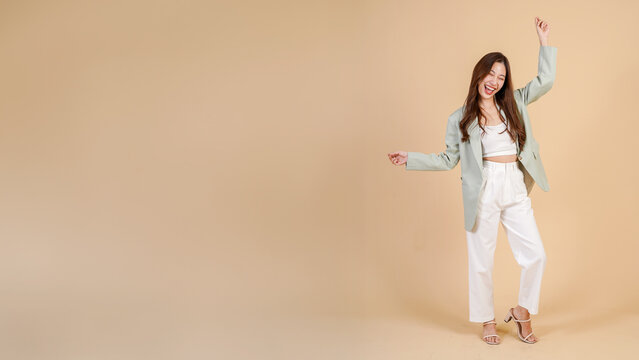 Young Asian happy smiling woman in casual business suit raising her hands up like a winner isolated over beige background - with empty copy space - in full body
