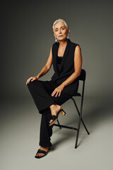 fashionable senior lady in black elegant attire sitting on chair on grey, classic fashion and age