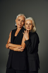 positive mature ladies in black stylish clothes looking at camera on grey, aging gracefully
