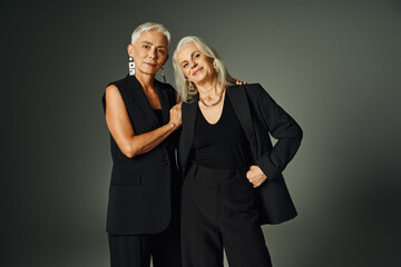happy senior model in black attire posing with hand on hip near elegant female friend on grey