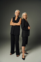 trendy senior lady in black attire posing with folded arms near female friend on grey, elegance