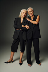full length of joyful senior models in black elegant clothes standing and smiling at camera on grey