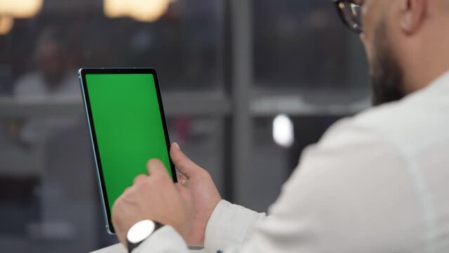 Bearded Man in Glasses Using Tablet Computer with Green Screen Mock Up Display. Male Watching Videos and Reading Social Media Posts on Mobile Device. Shooting Close Up Over the Shoulder