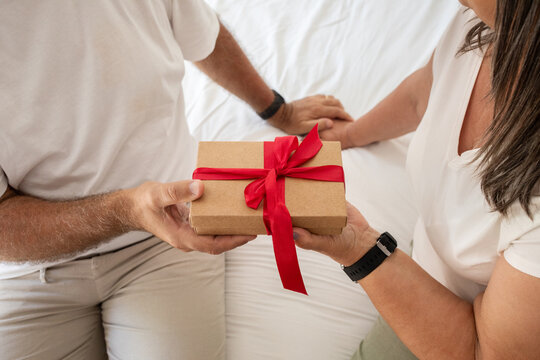 Positive senior caucasian woman gives gift box to man, sit on bad in bedroom interior - Powered by Adobe