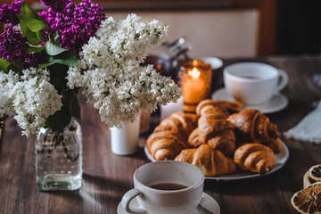 small croissants on a plate, a cozy hygge atmosphere, a glass teapot and cups of tea nearby. wooden background, lilac in a vase