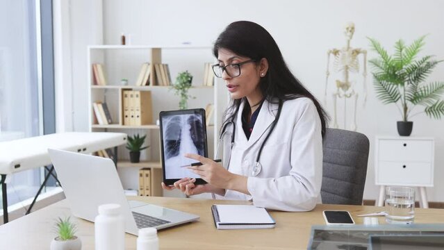 Concentrated female doctor talking on video call while showing result of examination of patient with tuberculosis. Indian lady consulting person using laptop and tablet in spacious clinic room.
