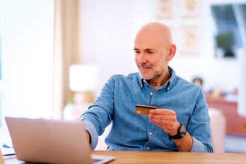 Confident mid aged man looking at bannk card and laptop while shopping online