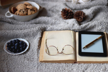 Bowl of cookies, cup of tea or coffee, plate of blueberries, dry oranges, wool yarn, pine cones, books, reading glasses, tablet, pen, soft blanket and lit candles on the table. Winter hygge at home.