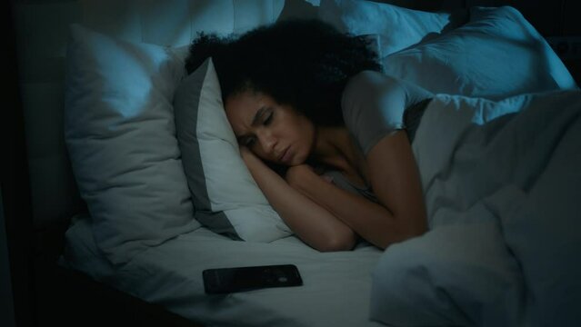 African American woman sleeping on bed waking up disturbed with alarm clock on phone at late night incoming call notification frustrated annoyed girl awake in nighttime turning off ringing smartphone