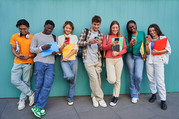 Obraz premium Cheerful multiracial group of young friends gathering to use phones while standing against a blue wall. Teenager students addicted to technology and social media looking at their own smartphones.