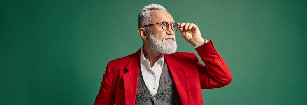 handsome Santa touching his glasses and looking away on green backdrop, winter concept, banner