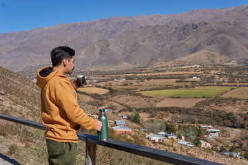 Fototapeta na wymiar latin man alone dressed in a mustard-colored sweatshirt, drinking mate looking at the landscape from a balcony in tafi del Valle, Tucuman, Argentina. Relaxed vacation
