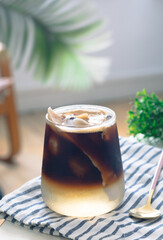 A clear glass of layer iced americano with young fresh coconut water and coconut meat on table with spoon and green leaves in background, home barista concept. 