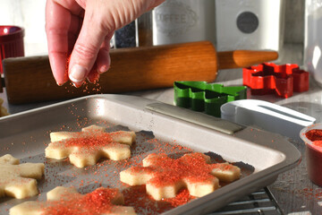 Sprinkling red sugar sprinkles onto Christmas cutout cookies dough on a baking sheet.