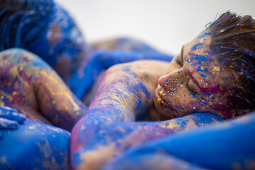 Young naked sexy woman in blue and magenta paint color painted, lies decorative sensual relaxed in elegant pose on the colorful studio floor. Creative, abstract expressive body art