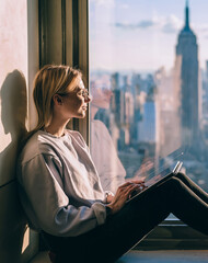 Young pensive female tourist inspired by scenery skyline of New York sitting on window