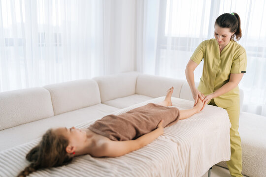 Wide shot of friendly female masseuse doing foot and legs massage to cute ten-year-old girl lying on massage table in spa salon. Little child having therapeutic, preventive massage session
