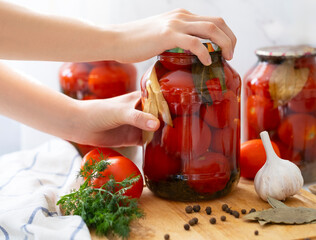 red tomatoes in canned jars stand on the kitchen table, female hands hold a jar nearby, spices and garlic