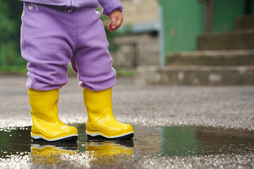 A happy  baby child in colorful clothes is jumping in a puddle. Fun on the street in summer. Splashes, drops of water in yellow waterproof boots jump in puddle and mud in the rain. 
