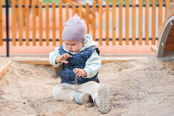 Cute baby kid sit in the sandbox and play with sand in a public park - obrazy, fototapety, plakaty