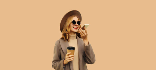 Beautiful smiling young woman with coffee cup holding cellphone using voice command recorder, assistant or takes calling wearing brown coat, round hat