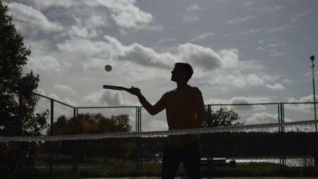 Silhouette of man hitting pickleball ball with racket on fenced court with net and serving. Player practicing hitting plastic ball with round holes during training outdoors in sunny weather.