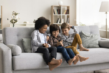Three multiethnic little boys spend time on internet sit together on sofa look at phone screen,...