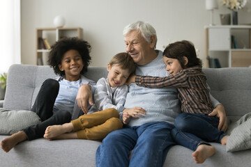 Three loving cute multiracial great-grandsons cuddling his grey-haired old great-grandpa express love and attachment sit together on couch enjoy time together at home. Multigenerational family ties - obrazy, fototapety, plakaty