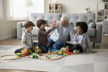 Great-grandpa gives high five to little great-grandson sit on floor spend pastime together with multiethnic great-grandchildren play wooden railroad, multi-generational family ties, games, playtime