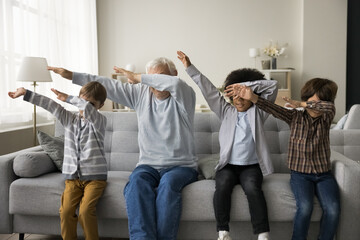 Older great-grandfather and three great-grandsons sit on couch in modern living room make dab popular youth culture gesture, multigenerational family perform triumph dance have fun together at home