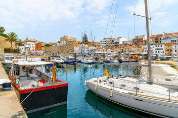 The picturesque old port harbor of the historic city of Ciutadella de Menorca, Spain, with fishing boats in the marina and the shops and cafes in view under the walled medieval city.	