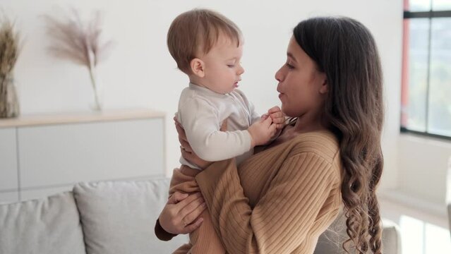 In the living room, a devoted mother holds little son in arms, showering him with tender kisses. Her eyes are filled with love, and the room is enveloped in the warmth of affectionate bond.