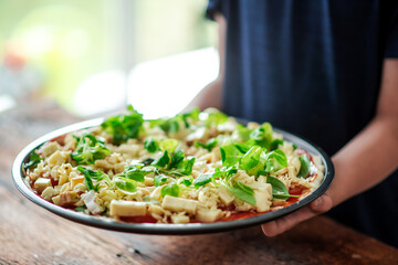 Raw pizza in a round shape with a lot of cheese and herbs in the hands of a child.