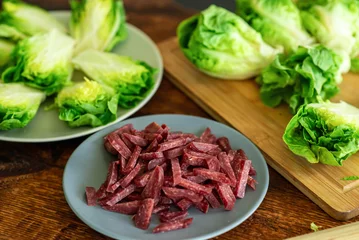 Fotobehang Fresh green lettuce cut into halves on a wooden cutting board. View from above. Chopped sausage. Recipe for stewed salad with meat. © Konstiantyn Zapylaie