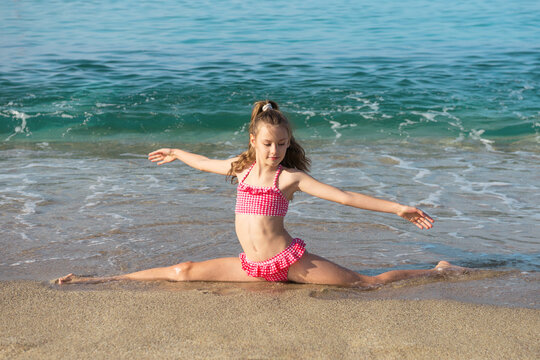 Pretty Little Girl In Red Swimming Suit Doing Splits On The Beach.