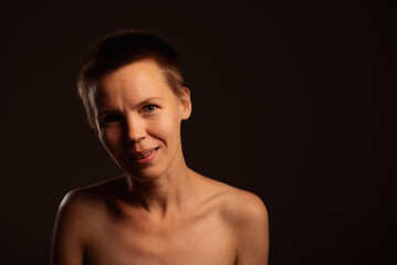 Captured against a black backdrop, this image portrays a middle-aged woman with a short haircut and exposed shoulders. Her enchanting face radiates a playful and inviting expression
