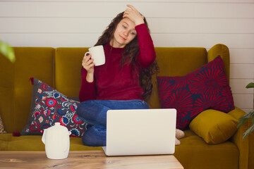 A young beautiful woman with long curly hair is relaxing at home sitting on a brown sofa with a...