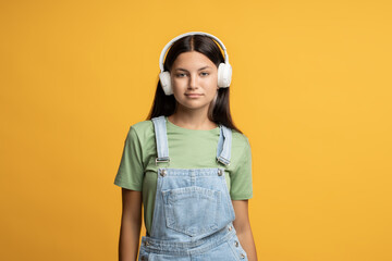 Girl music lover in headphones listening to music looking at camera on yellow background. Relaxed brunette teenager melomaniac enjoys favourite song earphones. Music fan hobby leisure pastime concept.