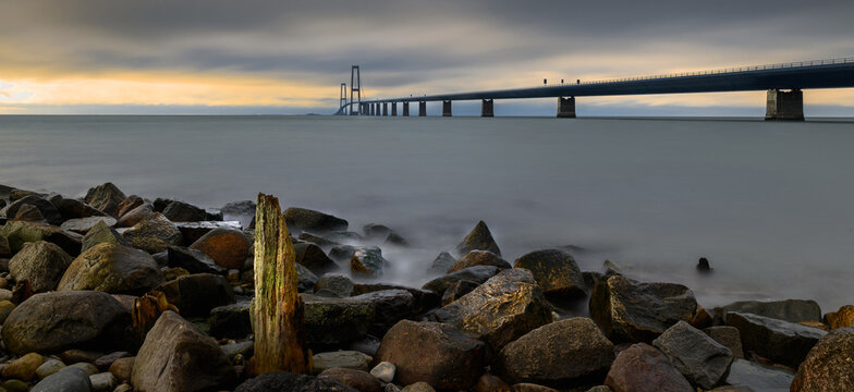 Large stones and weathered wooden stems as wavebreakers stand before the iconic Storeb&aelig;lt Bridge spanning the horizon. Storeb&aelig;ltsbroen Denmark's Great Belt Bridge connecting two coasts. 