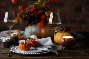 Elegant seasonal fall table setting with floral composition in a pumpkin vase as a centrepiece. White wine, burning candles, romantic cozy atmosphere of family dinner, private autumn wedding