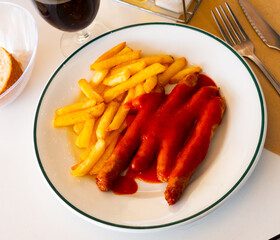 On white tablecloth of table there is plate with grilled sausages and slices of toasted potatoes