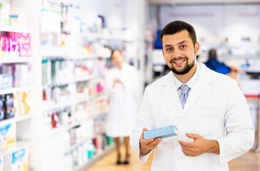 Portrait of european man druggist in lab coat standing in drugstore with drug package.