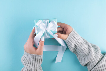 First person top view photo of female hands holding blue gift box with white ribbon. A pastel blue setting welcomes your words. my hands cradle a blue gift box