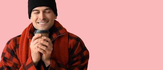 Happy young man in winter clothes and with cup of hot coffee on pink background