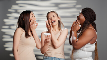 Group of flawless ladies applying moisturizing face cream in studio, promoting luminous skincare products. Interracial young women using moisturizer and serum for new dermatology campaign.