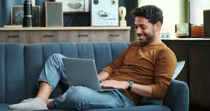Laid-back Middle Eastern man comfortably sits on a sofa with laptop. His relaxed posture highlights the ease and convenience of using technology for various purposes in a home setting.